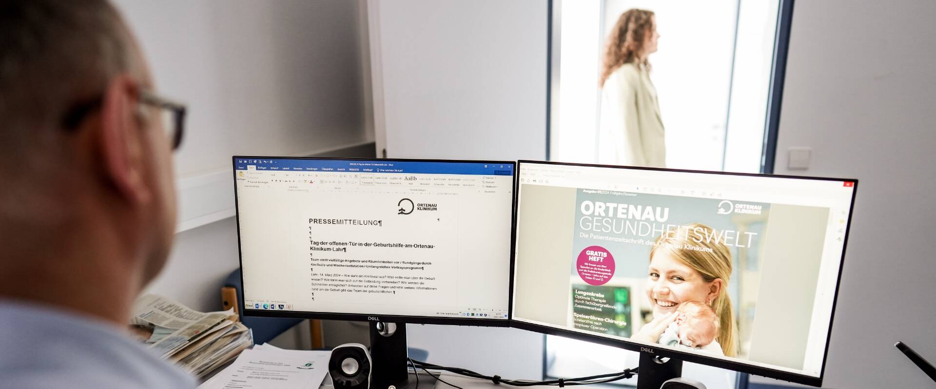 Blick auf zwei Monitore der Unternehmenskommunikation mit einem Mitarbeiter bei der Arbeit